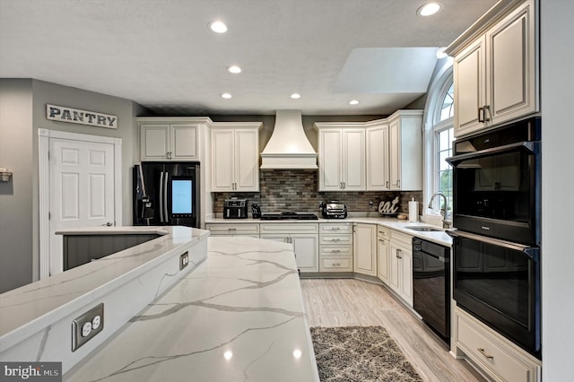 kitchen featuring light stone countertops, light hardwood / wood-style flooring, custom range hood, black appliances, and sink