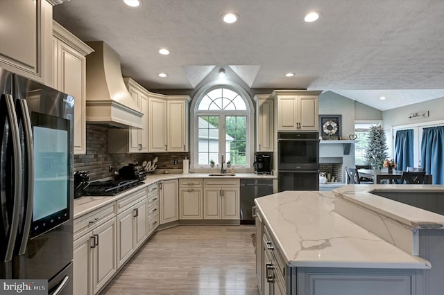 kitchen with cream cabinetry, black appliances, custom exhaust hood, light stone counters, and light hardwood / wood-style floors