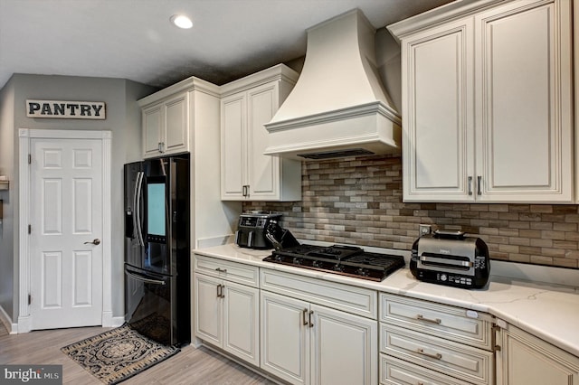 kitchen with decorative backsplash, light stone countertops, light wood-type flooring, custom range hood, and black appliances