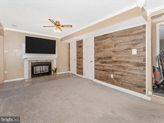 unfurnished living room with a tile fireplace, crown molding, wooden walls, carpet flooring, and ceiling fan