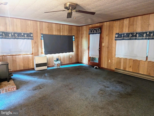 unfurnished living room with wood walls, baseboard heating, a wood stove, dark colored carpet, and ceiling fan