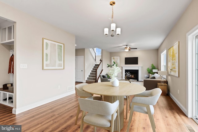 dining area featuring stairway, visible vents, a warm lit fireplace, ceiling fan with notable chandelier, and light wood-type flooring