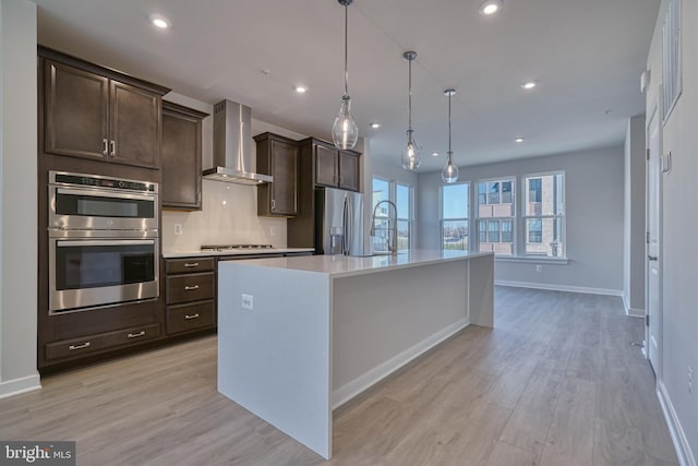 kitchen with appliances with stainless steel finishes, light hardwood / wood-style floors, decorative light fixtures, a center island with sink, and wall chimney range hood