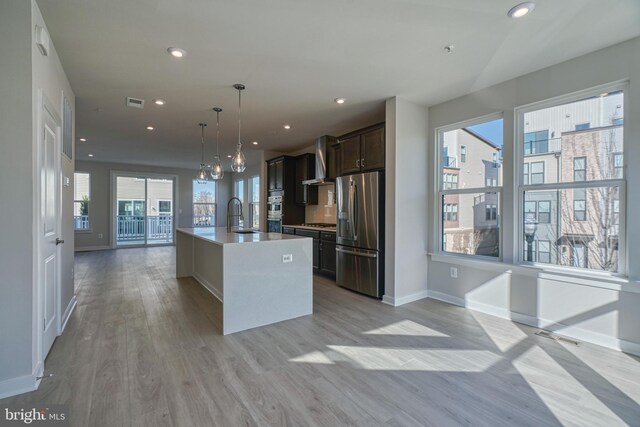 kitchen with appliances with stainless steel finishes, light wood-type flooring, decorative light fixtures, dark brown cabinetry, and a kitchen island with sink