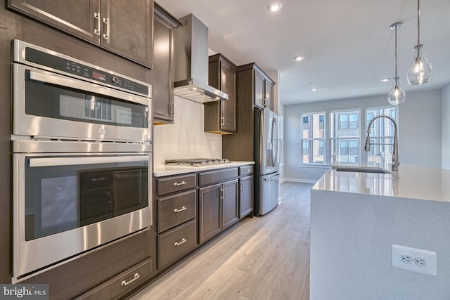 kitchen featuring light wood-type flooring, sink, hanging light fixtures, wall chimney exhaust hood, and appliances with stainless steel finishes