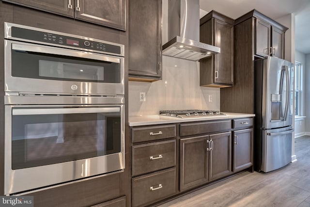 kitchen with appliances with stainless steel finishes, dark brown cabinetry, light wood-type flooring, and wall chimney range hood