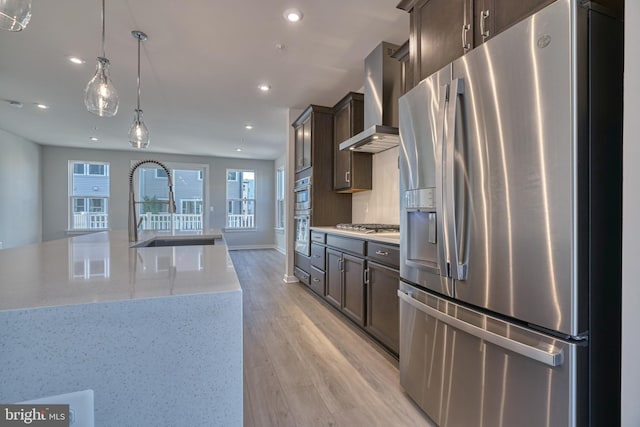 kitchen with hanging light fixtures, wall chimney exhaust hood, light stone countertops, stainless steel appliances, and light hardwood / wood-style flooring