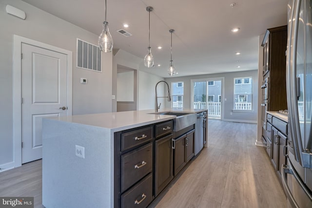 kitchen featuring hanging light fixtures, sink, a kitchen island with sink, appliances with stainless steel finishes, and light wood-type flooring