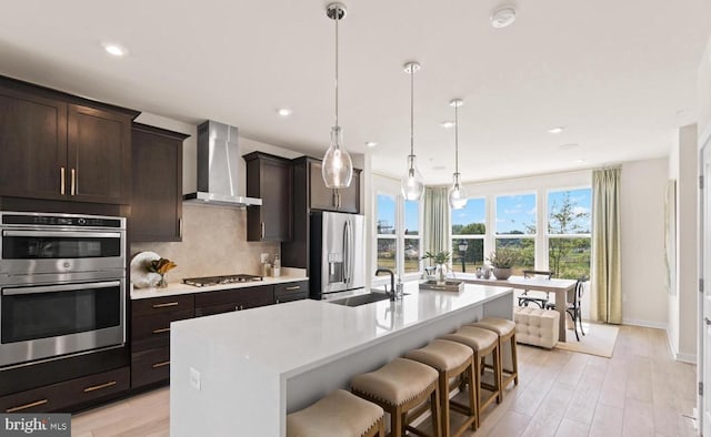 kitchen with sink, a kitchen island with sink, wall chimney range hood, appliances with stainless steel finishes, and a breakfast bar