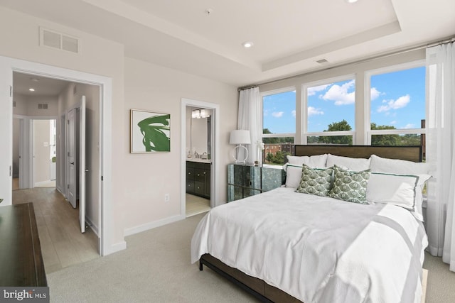 bedroom with multiple windows, a raised ceiling, ensuite bath, and light hardwood / wood-style flooring