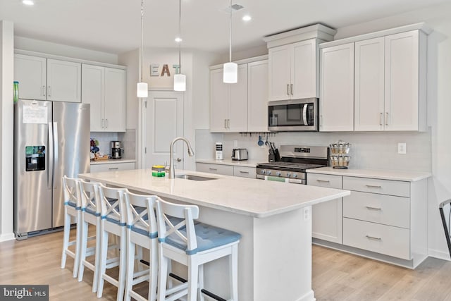 kitchen featuring light wood-type flooring, a kitchen island with sink, white cabinetry, stainless steel appliances, and decorative light fixtures