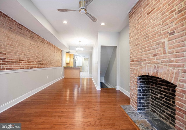 unfurnished living room featuring a brick fireplace, ceiling fan, brick wall, and hardwood / wood-style floors