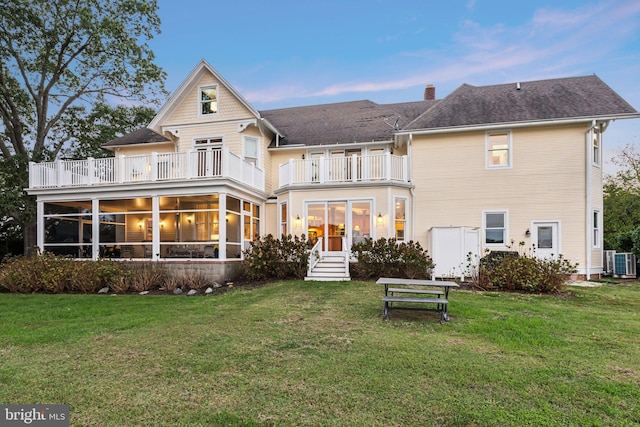 back house at dusk featuring a lawn, cooling unit, a balcony, and a sunroom