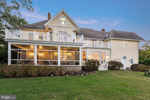 back house at dusk featuring a lawn, a balcony, and a sunroom