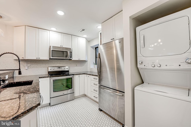 kitchen featuring white cabinets, stacked washer and clothes dryer, stainless steel appliances, dark stone counters, and sink