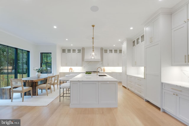 kitchen featuring hanging light fixtures, white cabinets, light wood-type flooring, a kitchen island with sink, and sink