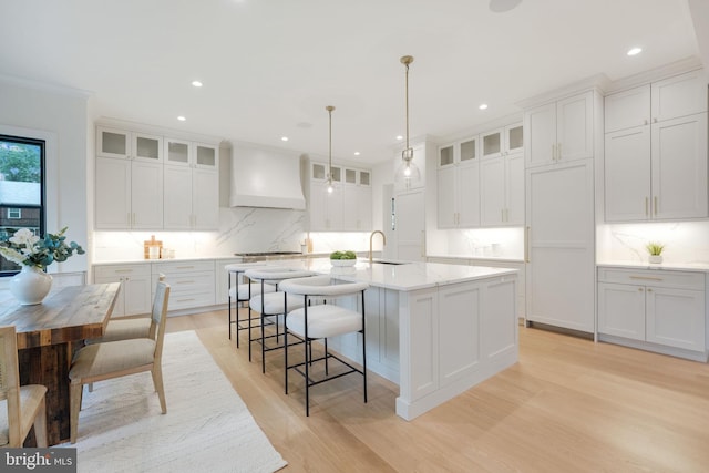 kitchen with white cabinets, decorative light fixtures, a kitchen island with sink, and premium range hood