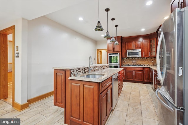 kitchen featuring stainless steel appliances, sink, light stone countertops, hanging light fixtures, and an island with sink