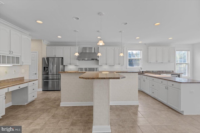 kitchen with wall chimney exhaust hood, a wealth of natural light, stainless steel refrigerator with ice dispenser, and a kitchen island