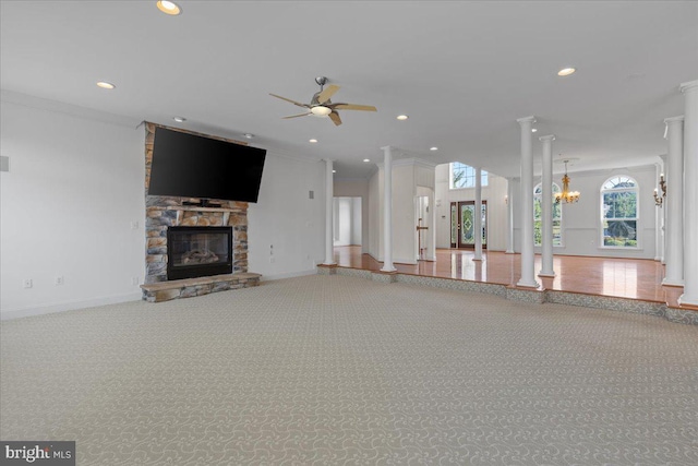 unfurnished living room featuring ceiling fan with notable chandelier, a stone fireplace, crown molding, carpet flooring, and decorative columns