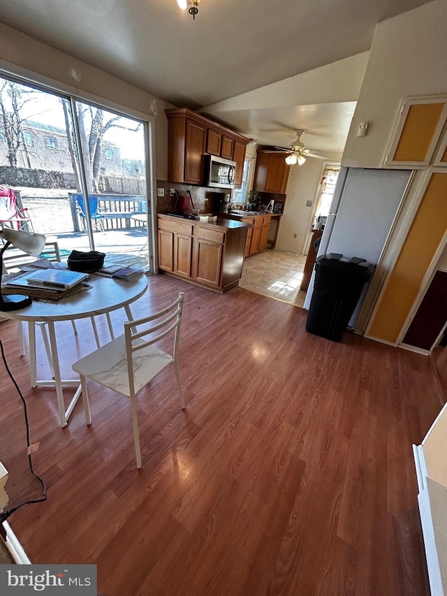 kitchen with brown cabinetry, ceiling fan, stainless steel microwave, wood finished floors, and vaulted ceiling