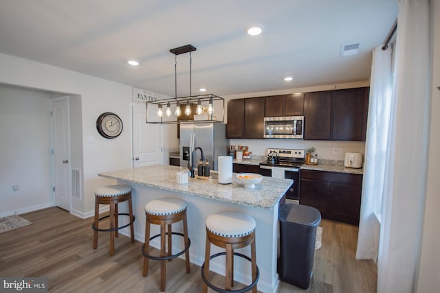 kitchen featuring a kitchen island with sink, appliances with stainless steel finishes, hanging light fixtures, and light hardwood / wood-style flooring