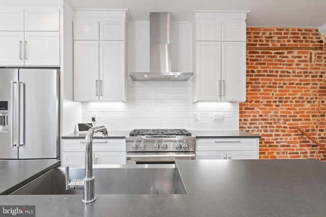 kitchen featuring wall chimney range hood, tasteful backsplash, white cabinetry, premium appliances, and brick wall