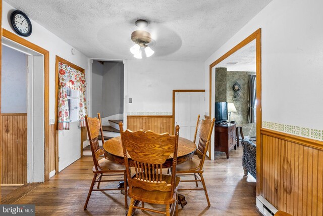 dining space featuring a textured ceiling, ceiling fan, hardwood / wood-style flooring, wooden walls, and a baseboard heating unit