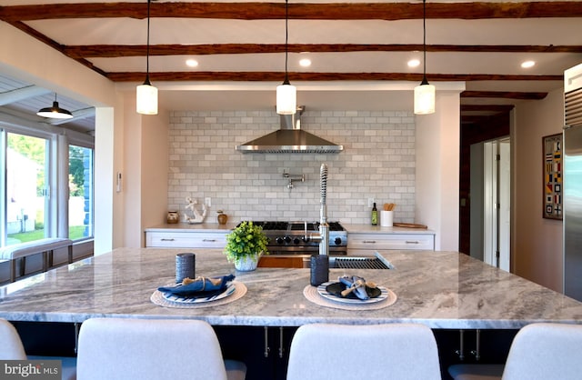 kitchen with light stone counters, backsplash, wall chimney range hood, decorative light fixtures, and a spacious island