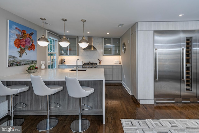 kitchen featuring sink, built in refrigerator, decorative light fixtures, wall chimney range hood, and dark hardwood / wood-style flooring