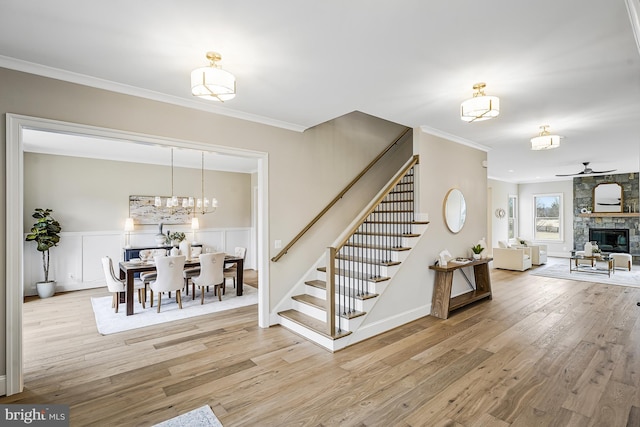 interior space with a stone fireplace, ceiling fan with notable chandelier, wood finished floors, wainscoting, and crown molding