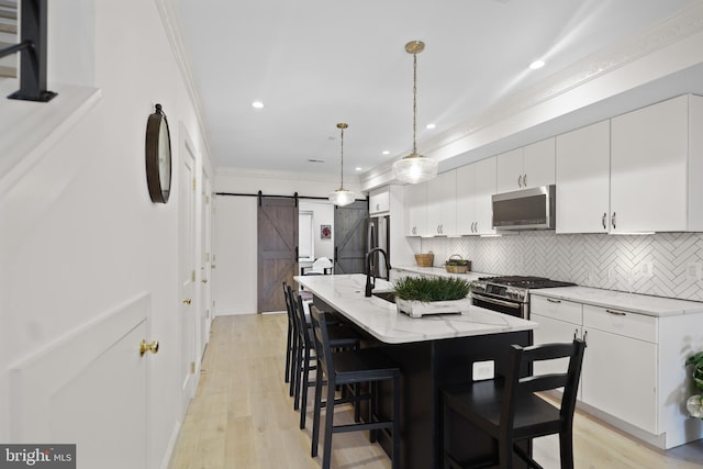 kitchen featuring white cabinets, an island with sink, decorative light fixtures, stainless steel appliances, and a barn door