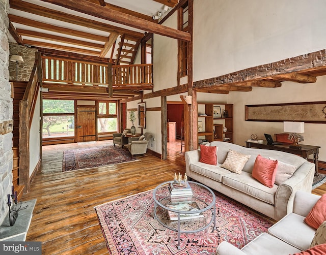 living room featuring wood-type flooring, a stone fireplace, beamed ceiling, and a towering ceiling