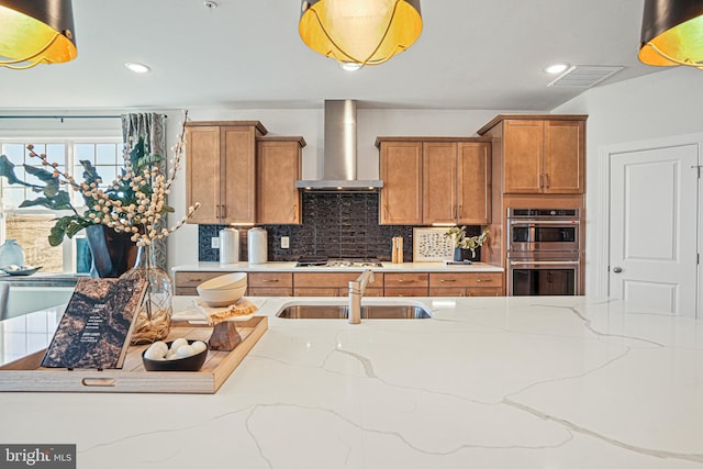 kitchen featuring decorative backsplash, light stone counters, wall chimney exhaust hood, stainless steel double oven, and sink