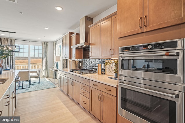 kitchen with wall chimney range hood, hanging light fixtures, light hardwood / wood-style flooring, light stone countertops, and appliances with stainless steel finishes