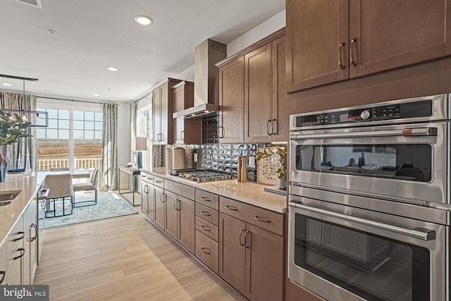 kitchen with light stone countertops, wall chimney range hood, light hardwood / wood-style floors, decorative light fixtures, and appliances with stainless steel finishes