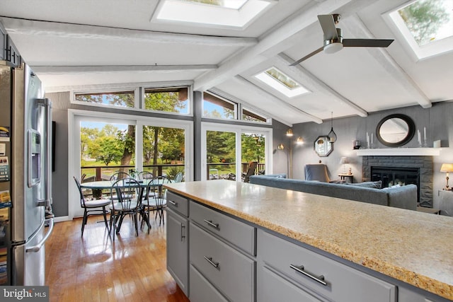 kitchen with light wood-type flooring, ceiling fan, lofted ceiling with skylight, a stone fireplace, and stainless steel refrigerator with ice dispenser