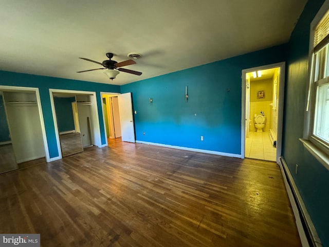 unfurnished bedroom featuring dark hardwood / wood-style floors, baseboard heating, ensuite bath, two closets, and ceiling fan