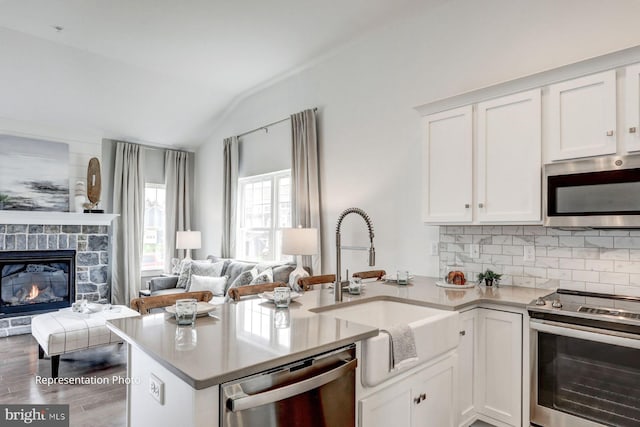 kitchen with appliances with stainless steel finishes, vaulted ceiling, white cabinets, a stone fireplace, and hardwood / wood-style floors