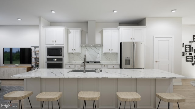 kitchen with appliances with stainless steel finishes, white cabinetry, wall chimney exhaust hood, a breakfast bar area, and a kitchen island with sink