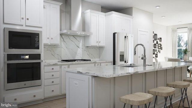 kitchen featuring tasteful backsplash, a kitchen island with sink, wall chimney range hood, appliances with stainless steel finishes, and a breakfast bar