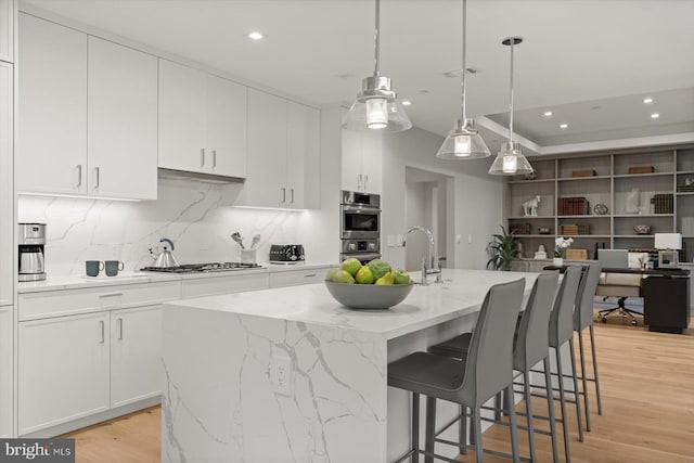 kitchen featuring white cabinets, an island with sink, decorative light fixtures, appliances with stainless steel finishes, and light wood-type flooring