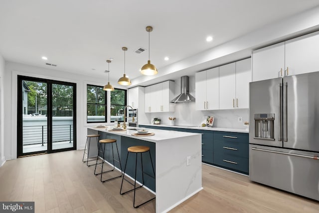 kitchen featuring white cabinets, pendant lighting, stainless steel appliances, a kitchen island with sink, and wall chimney range hood