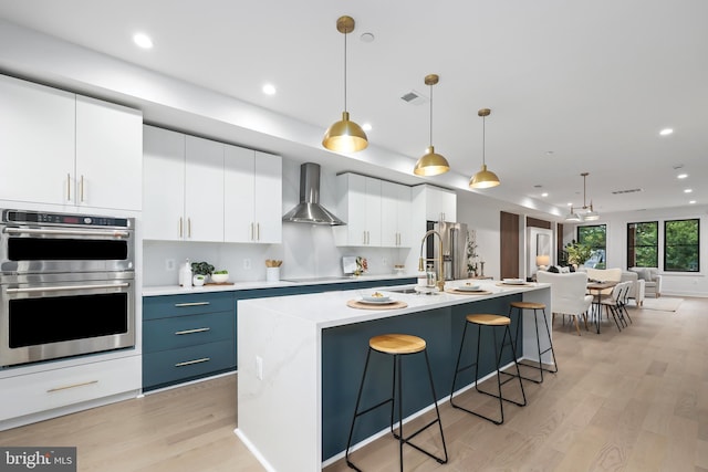 kitchen featuring pendant lighting, double oven, wall chimney exhaust hood, and a kitchen island with sink