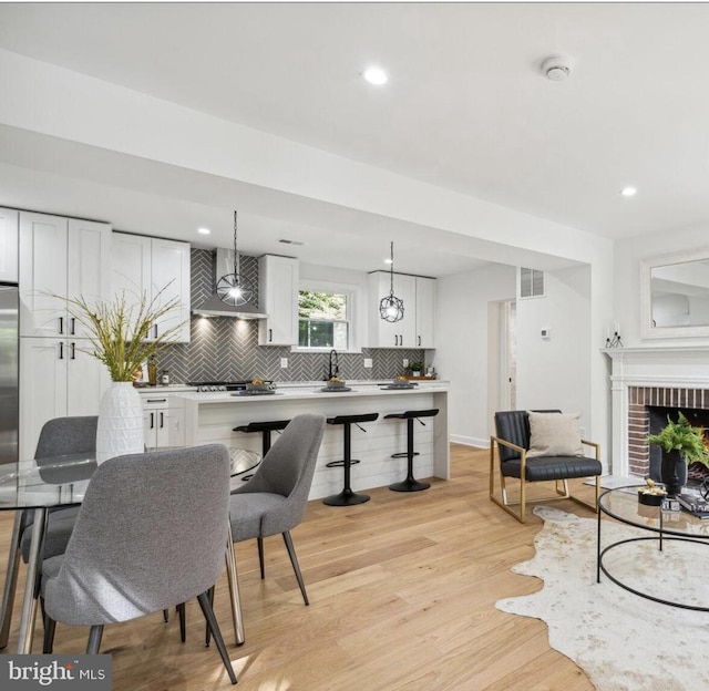 kitchen with light wood-type flooring, white cabinetry, hanging light fixtures, wall chimney exhaust hood, and a brick fireplace