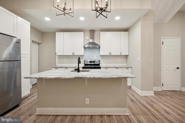 kitchen featuring appliances with stainless steel finishes, wall chimney exhaust hood, an island with sink, and decorative light fixtures