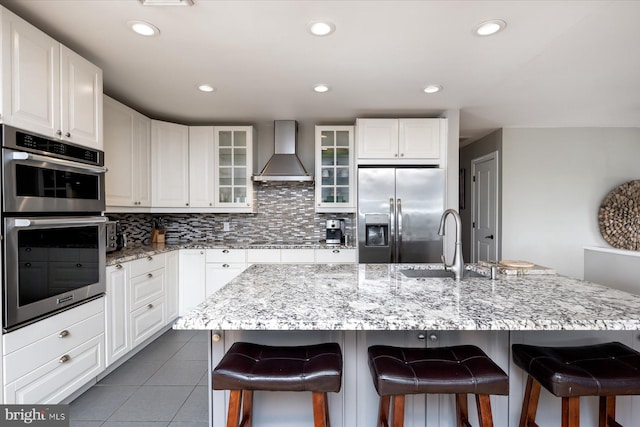 kitchen featuring wall chimney exhaust hood, sink, a center island with sink, appliances with stainless steel finishes, and white cabinets