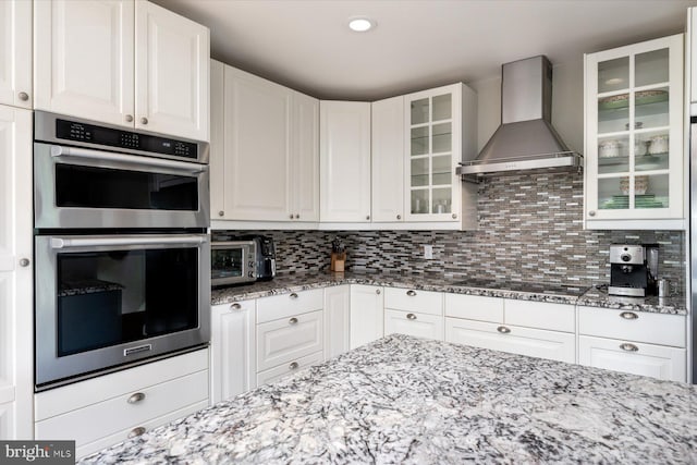 kitchen featuring stainless steel double oven, stovetop, light stone countertops, wall chimney exhaust hood, and white cabinetry