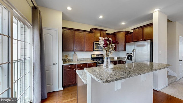 kitchen featuring a breakfast bar, light stone counters, hardwood / wood-style flooring, a center island with sink, and appliances with stainless steel finishes