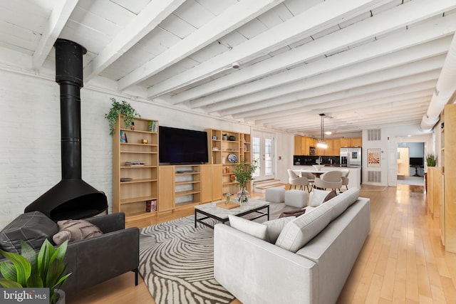 living room featuring a wood stove, light hardwood / wood-style flooring, beamed ceiling, and brick wall